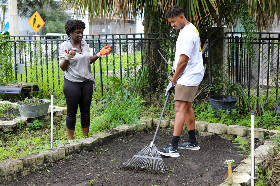 Two people gardening: a woman explaining with orange gloves while a man rakes soil in a raised garden bed.
