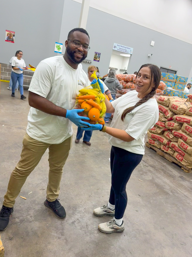 Two volunteers in white shirts exchange a bag of mixed fruit (bananas and oranges) in a donation warehouse with stacks of produce in the background.