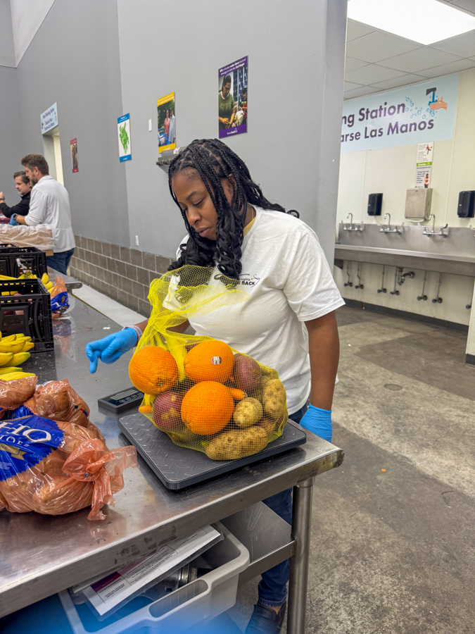 Volunteer wearing a white shirt and gloves weighs a mesh bag of fruit on a scale at a food pantry table.
