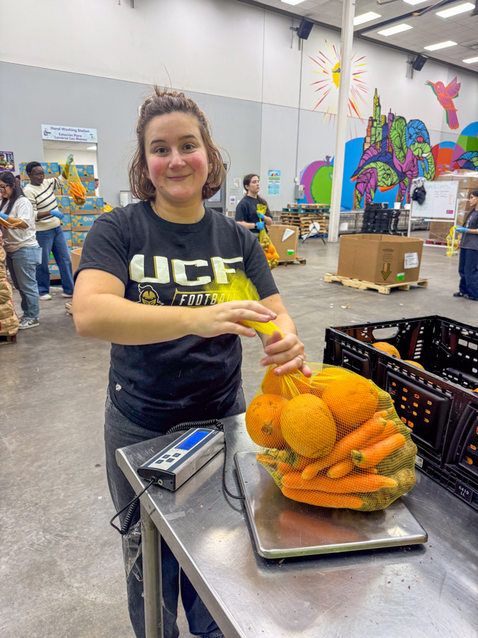 Woman weighs a mesh bag of oranges and carrots on a scale at a produce distribution center, with volunteers in the background.