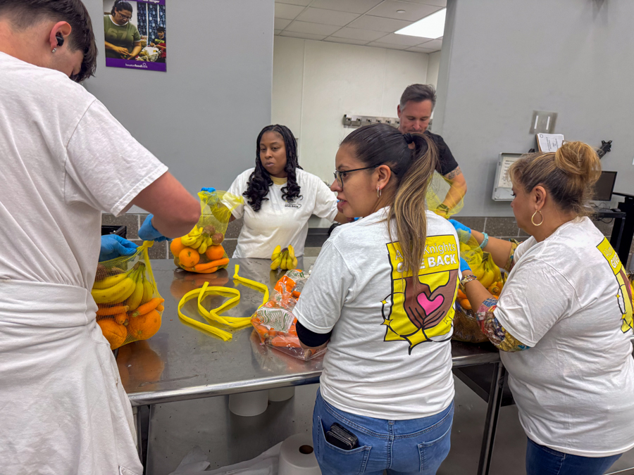 Group of volunteers packing fruit into yellow bags at a stainless steel table in a kitchen or food bank setting, preparing donations.