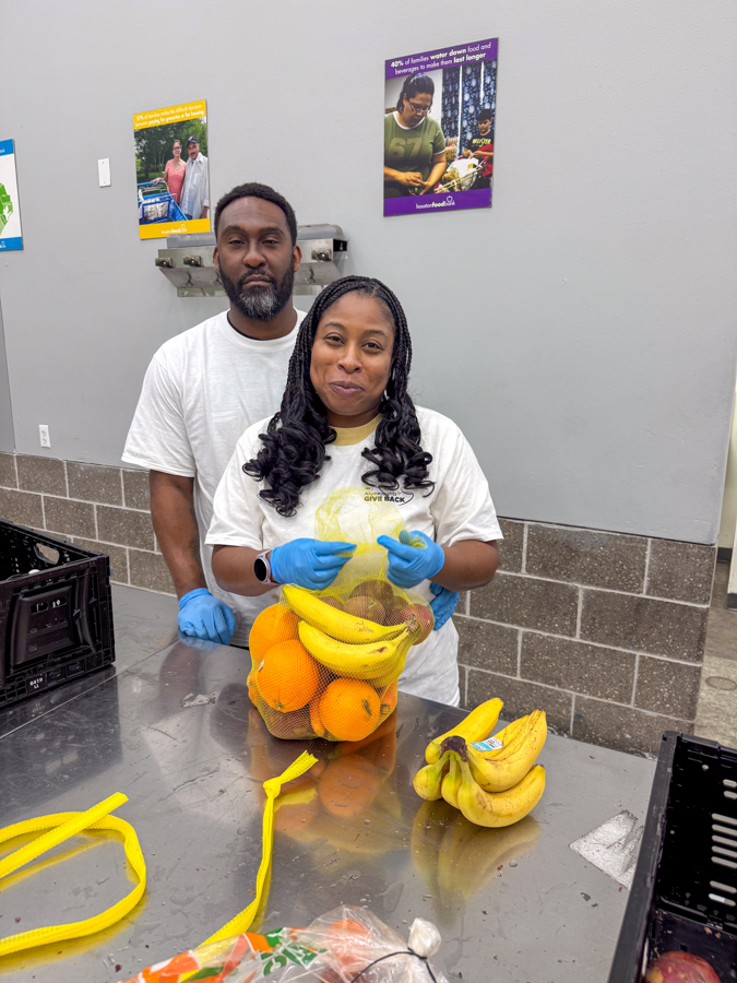 Two volunteers package fruit at a donation station: a smiling woman in front places bananas and oranges into a mesh bag while a man stands behind her, both wearing gloves and white shirts on a stainless table.