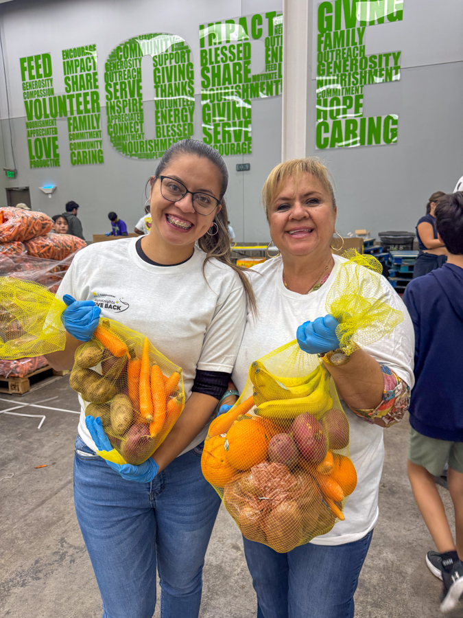 Two smiling volunteers in white shirts and blue gloves hold mesh bags of carrots, potatoes, and other produce at a food bank charity event.