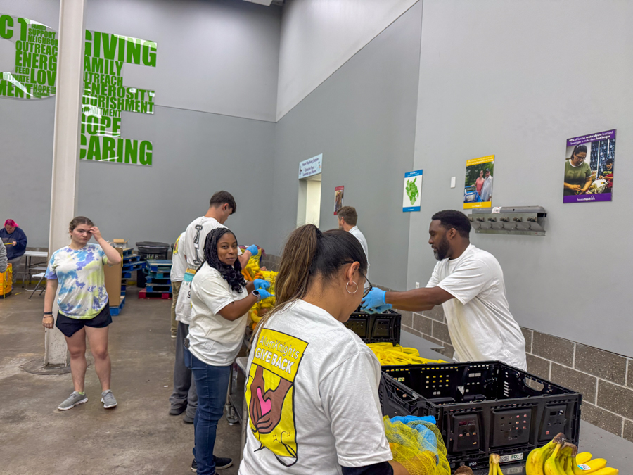 Volunteers in a donation center packing yellow bags of produce on a table with crates of bananas nearby.