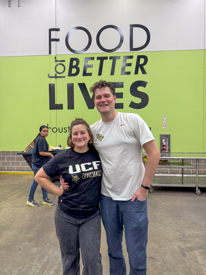 Two smiling people pose with their arms around in front of a green wall banner that reads 'FOOD for BETTER LIVES'; a person serves food in the background.