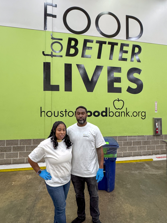 Two volunteers in white shirts and blue gloves stand together in front of a bright green wall that reads 'FOOD for BETTER LIVES' and 'houstonfoodbank.org'.