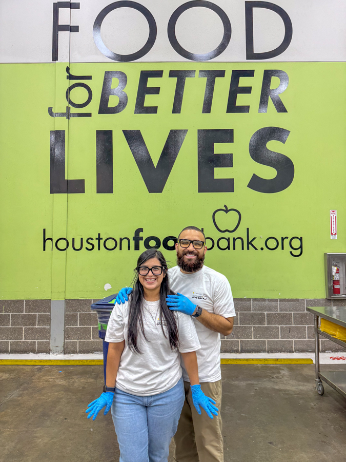 Two volunteers wearing white shirts and blue gloves stand smiling in front of a lime green Houston Food Bank mural that reads 'FOOD for BETTER LIVES' (houstonfoodbank.org).