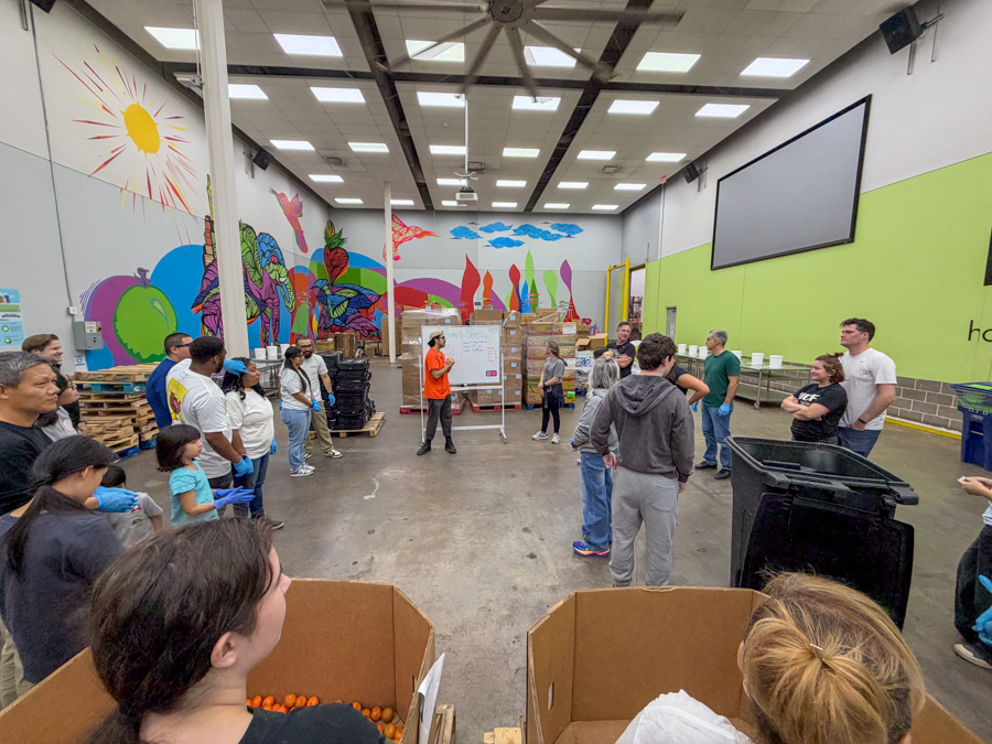 Group of people standing in a warehouse listening to a presenter in an orange shirt at a whiteboard, with a colorful mural on the walls and a large screen overhead.