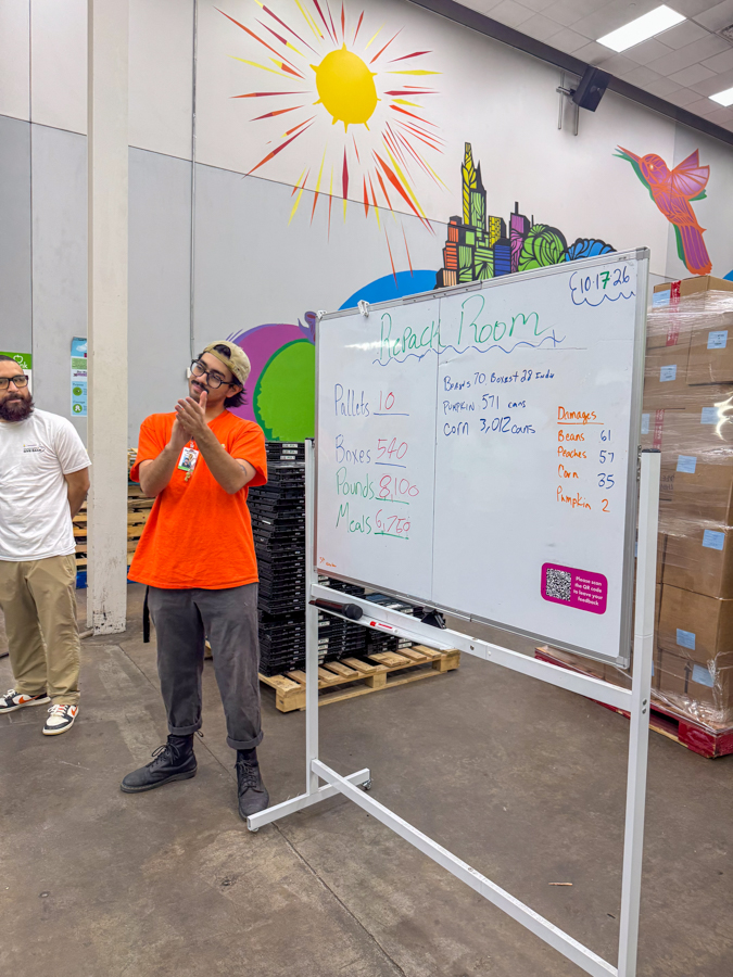 Person in an orange shirt presents beside a whiteboard in a warehouse with a colorful mural; board shows tallies and ‘Beach Room’ heading.