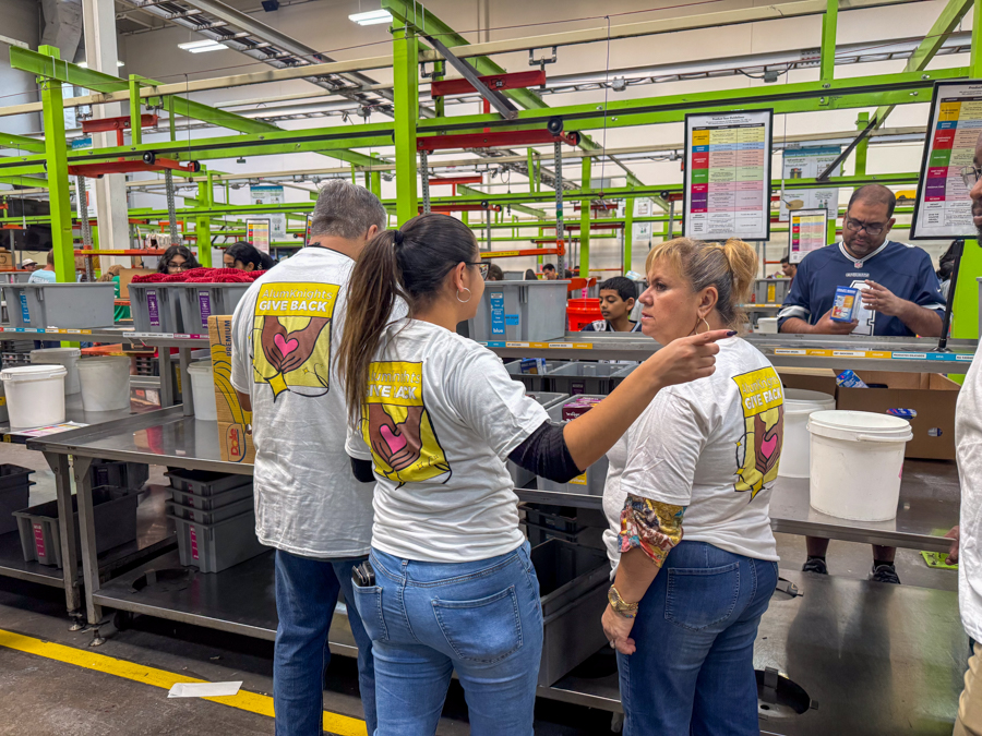 Group of volunteers in white T-shirts with a 'Give Back' logo, assisting at a production line in a bright factory setting.