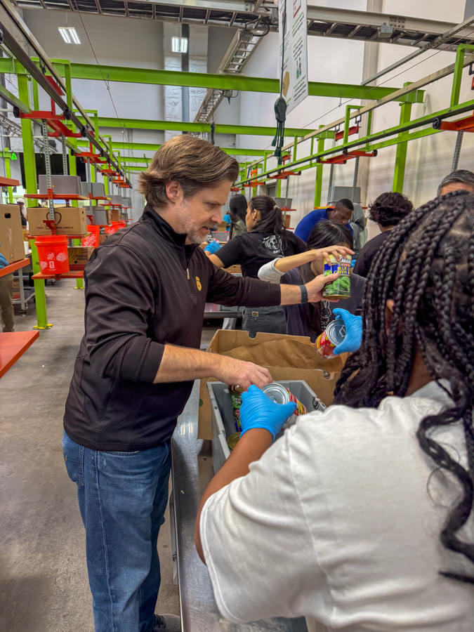 Volunteers in a distribution center sort and pack canned foods at a long worktable, wearing gloves.