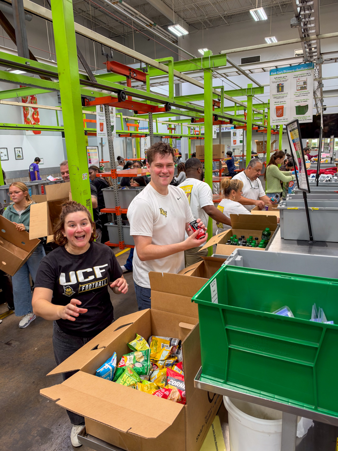Group of volunteers packing snacks at a warehouse donation event; green metal racks, open boxes of chips, and people sorting items with smiling faces in the foreground.