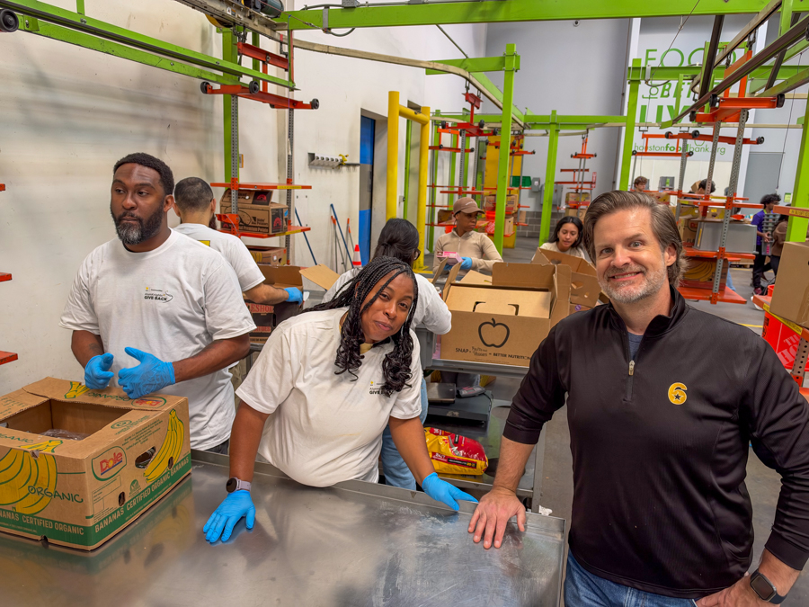 Volunteers in white shirts and blue gloves packing boxes at a busy food bank with green shelving in the background.