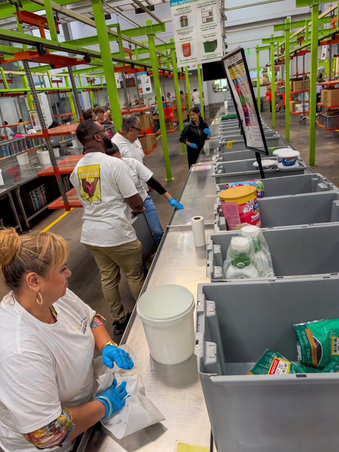 Workers wearing blue gloves sort items into gray bins along a stainless sorting counter in a bright warehouse with green support beams.