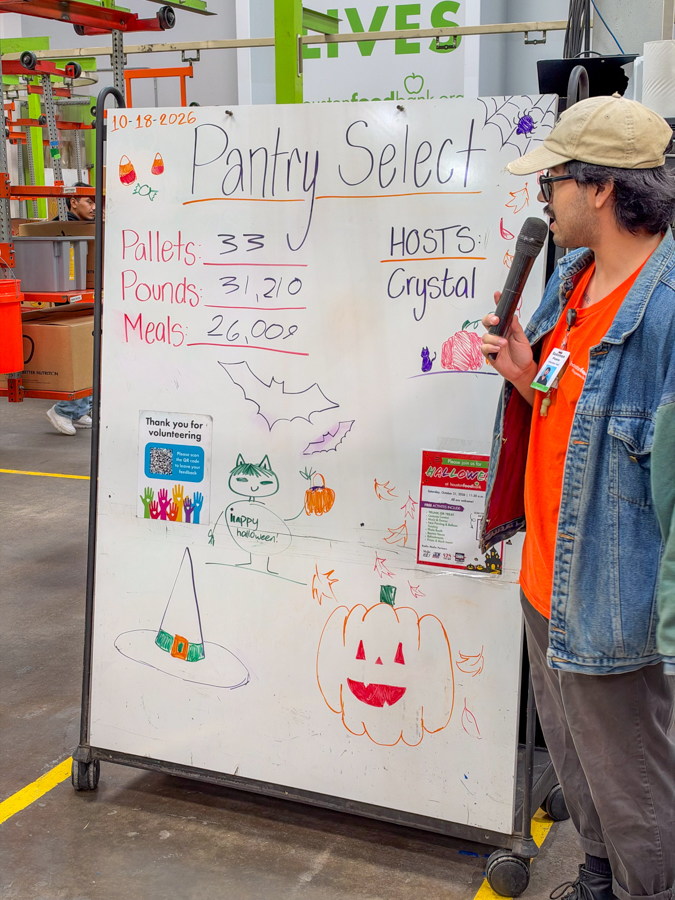 Person in a cap and orange shirt speaks into a microphone beside a whiteboard reading 'Pantry Select' with numbers and drawings.