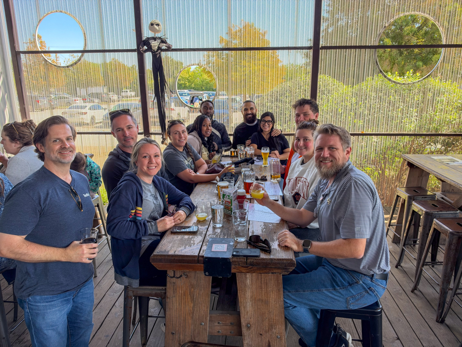A group of about twelve people sits around a long wooden table on an outdoor patio, smiling and enjoying drinks together.