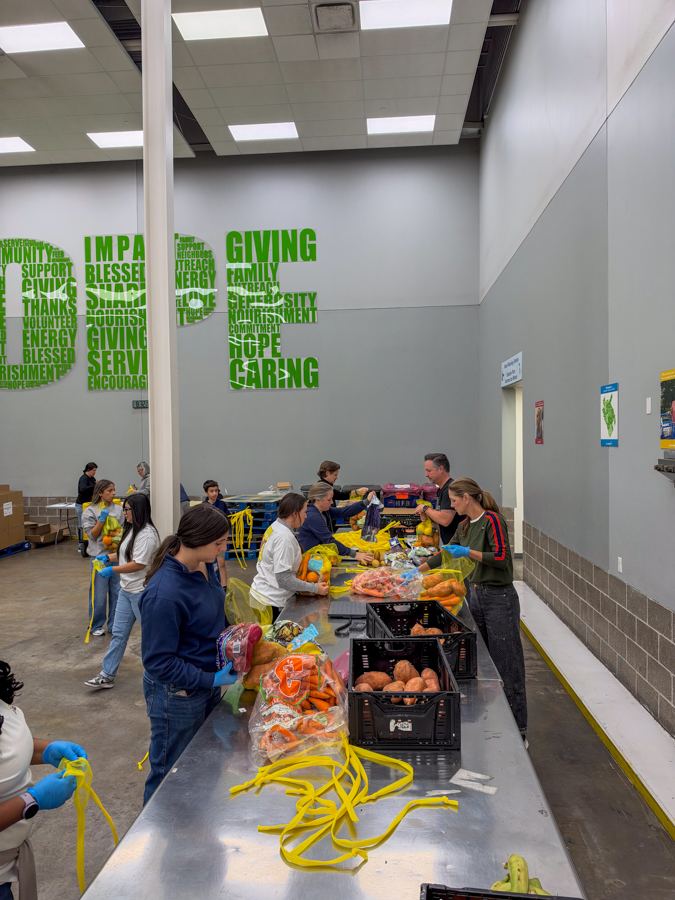 Volunteers at a long table packing produce into bags during a community food drive, with pumpkins and yellow straps nearby under a 'GIVING' mural in the background