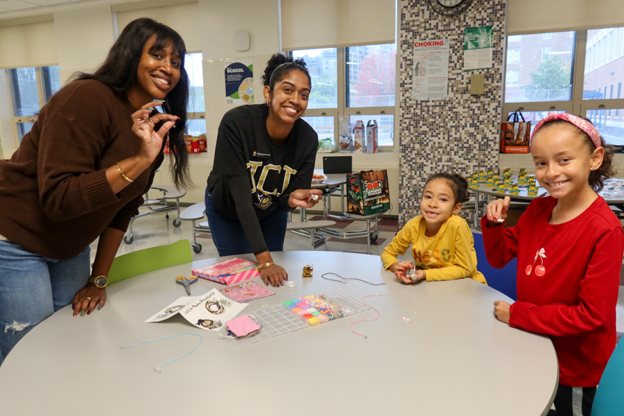 Four people—two adults and two children—smile at a round table in a bright classroom while making bead crafts with colorful supplies laid out before them.