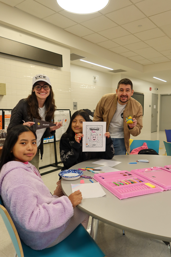 Four students sit and stand around a round table in a bright room, smiling; art supplies and a coloring page are on the table.
