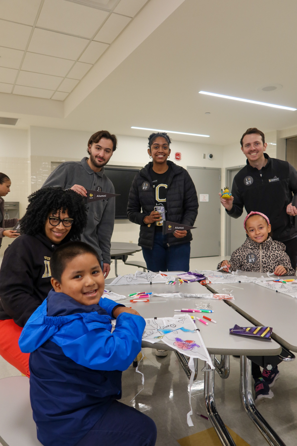Group of students and volunteers smile at a classroom craft activity around a table with markers and drawings on paper.