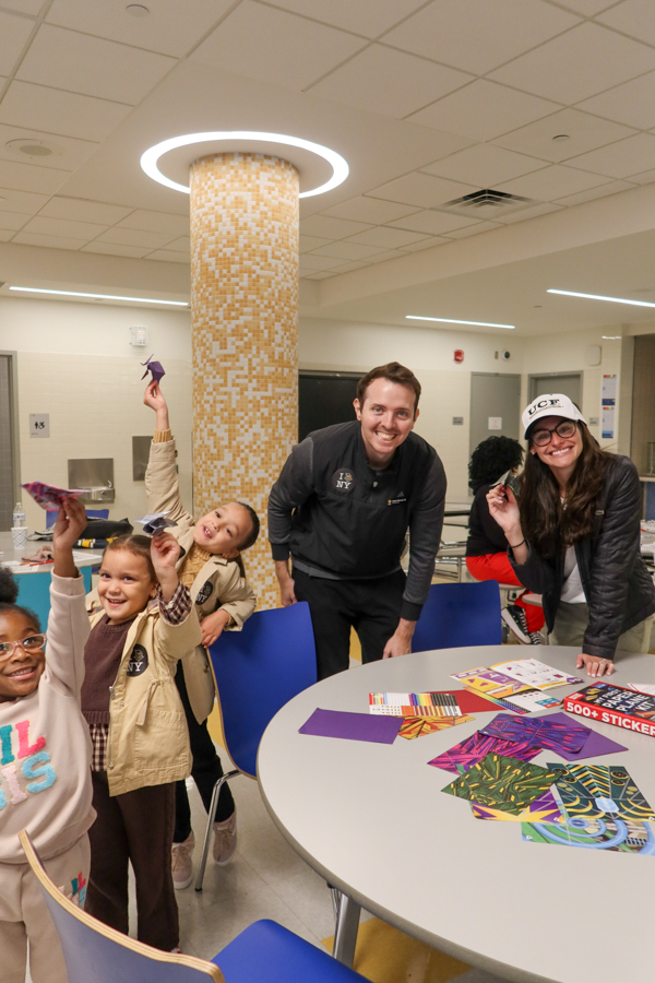 Group of children and two adults at a round table with colorful stickers, smiling during a craft activity in a bright room.