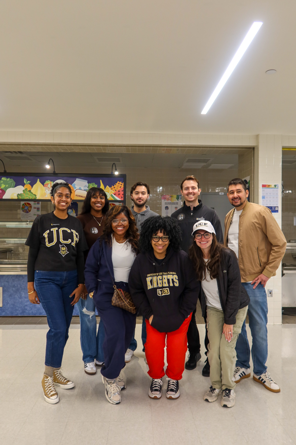 Group of eight smiling students posing in a school cafeteria with a lunch counter in the background.