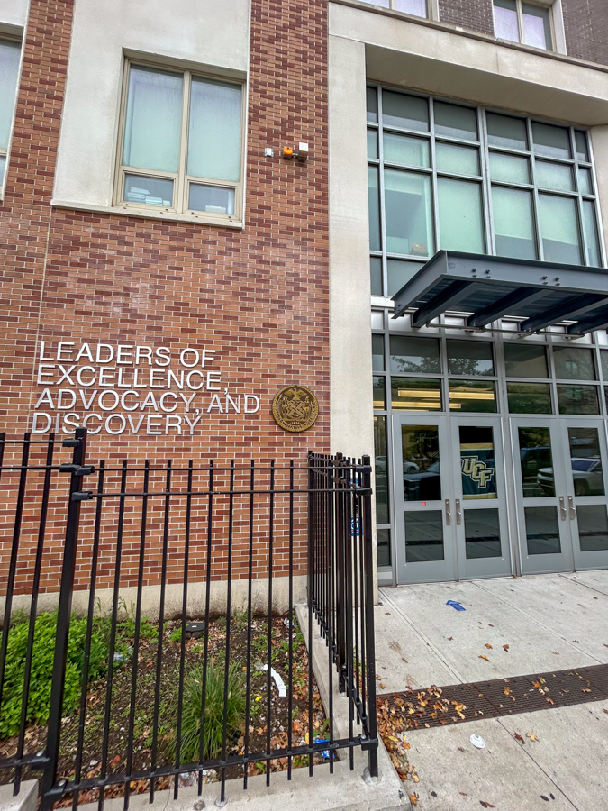 Brick campus building entrance with glass doors, a metal fence, and a sign: 'Leaders of Excellence, Advocacy, and Discovery' near a round emblem on the brick wall.