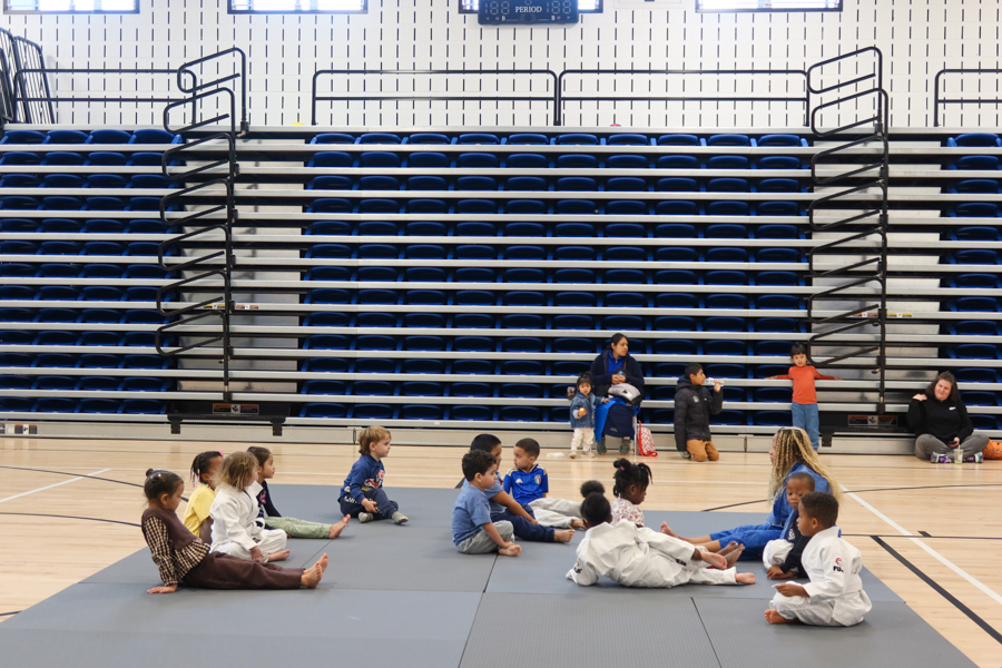 Group of young children in martial arts uniforms sitting on gray mats in a gym, with blue bleachers in the background and a few adults watching from the sidelines.
