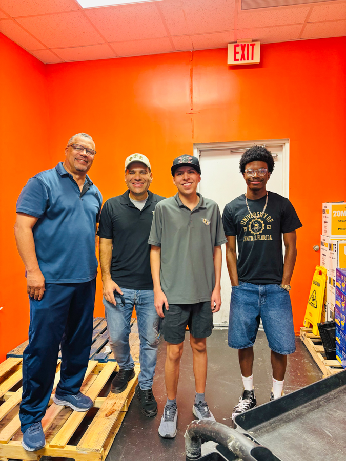 Four men stand side by side in a bright orange room, smiling at the camera.