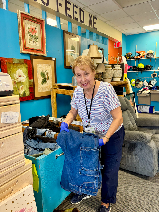 Smiling female thrift-store volunteer holds a folded denim jacket, wearing gloves and a name badge in a bright blue shop.