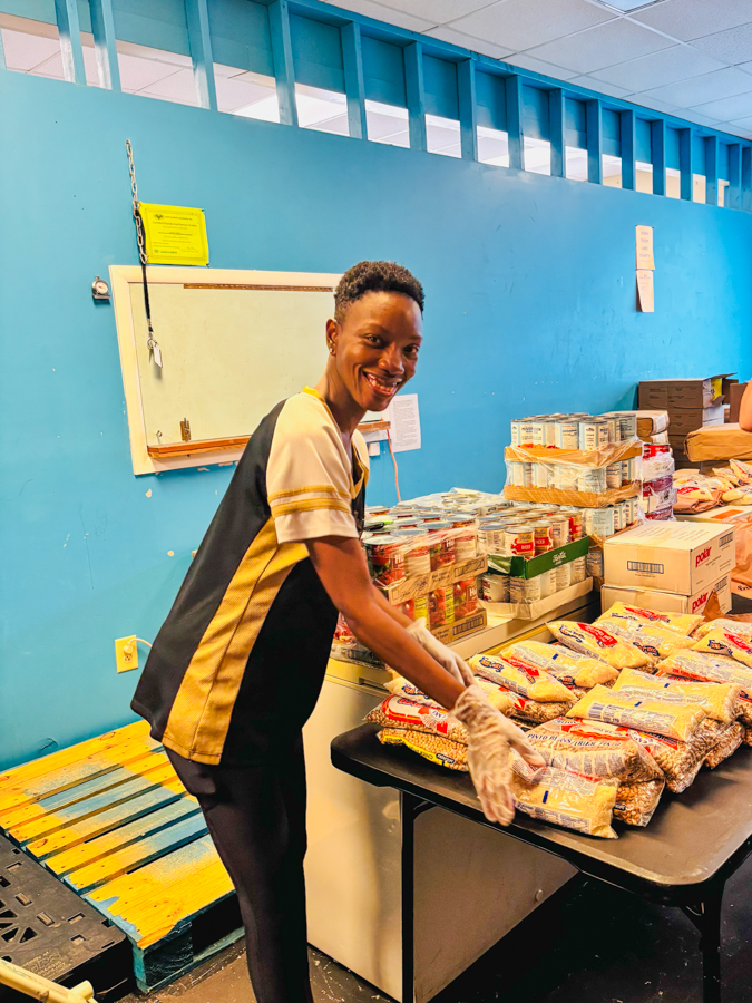 Volunteer smiling while packing bags of food on a table in a blue-walled room.