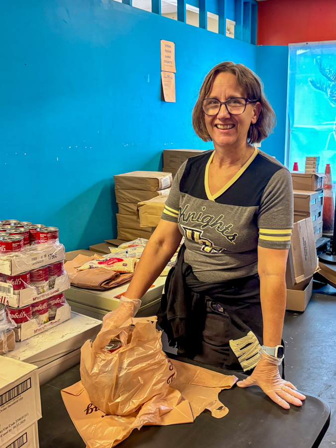 Smiling woman wearing gloves packs groceries into a brown paper bag at a food bank, with canned goods and boxes in the background on a bright blue wall.