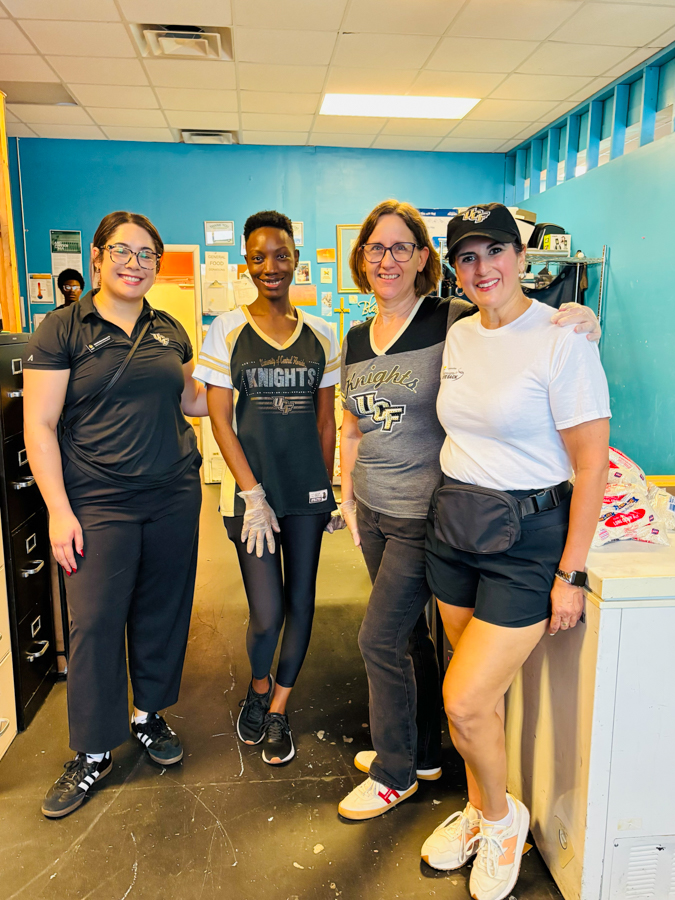 Four women stand together in a blue community room, smiling at the camera. Left to right: woman in black polo, girl in a Knights jersey with gloves, woman in a gray Knights shirt, and woman in a white tee with cap and waist pack.