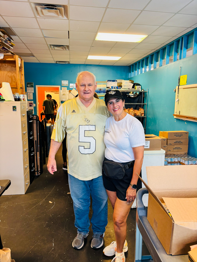 Two volunteers stand together in a blue-walled storage room, smiling at the camera; man wears a gold Florida Gators jersey with number 5, jeans, and sneakers while the woman wears a white t-shirt, cap, and a waist pouch.