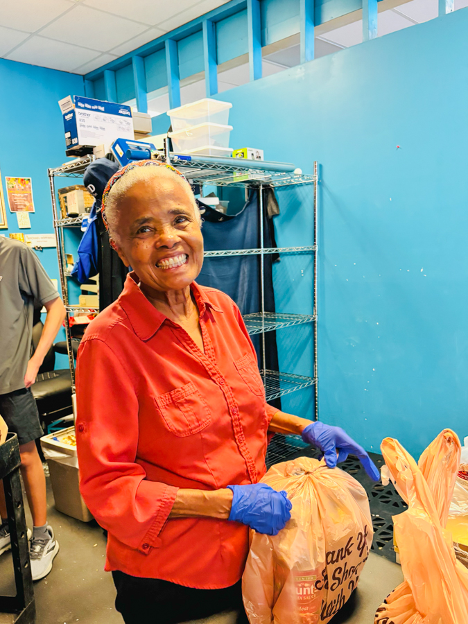 Smiling woman in a red shirt with blue gloves packs groceries into a plastic bag in a storage room with metal shelves behind her.