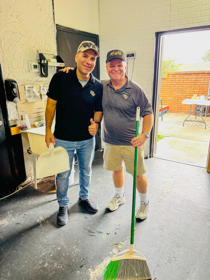 Two men stand inside a workshop, one holding a broom and giving a thumbs up, smiling at the camera.