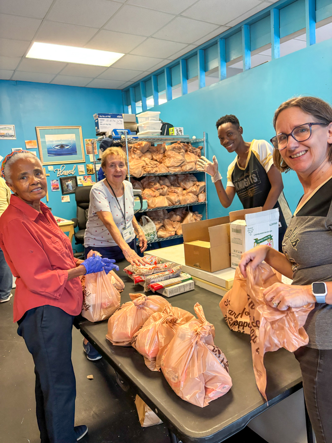 Four volunteers pack grocery bags on a table in a blue-walled pantry, with bread on shelves in the background.