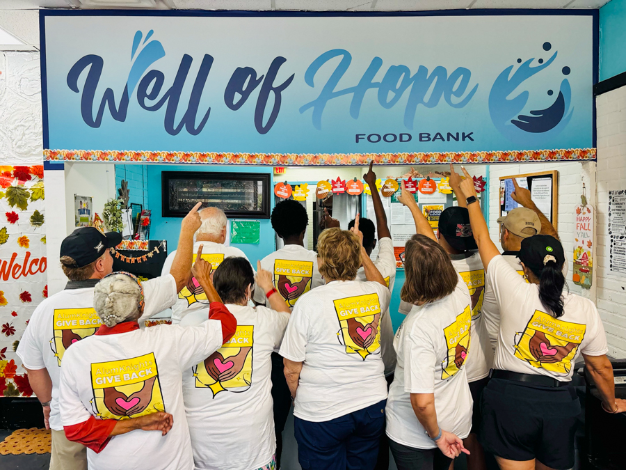 Group of volunteers in white shirts with yellow logos raise their hands inside a Food Bank under a blue 'Well of Hope' sign on the wall, celebrating together.