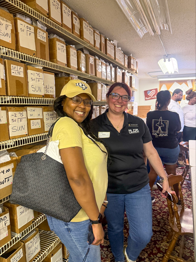 Two women pose for a photo in a storage room with labeled boxes on metal shelves; one wears a yellow shirt and cap, the other a black polo shirt, both smiling.