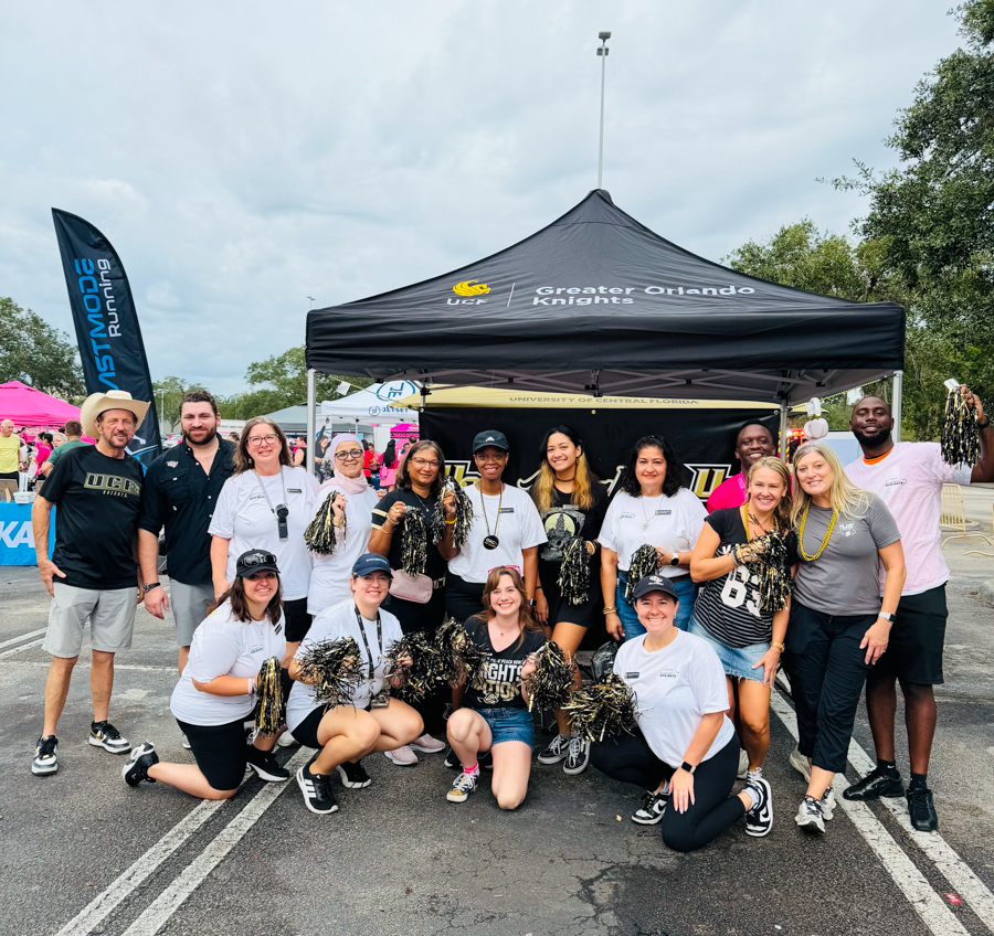 Group of volunteers and athletes posing with gold/black pom-poms in front of a black tent labeled 'Greater Orlando Knights' at an outdoor event.
