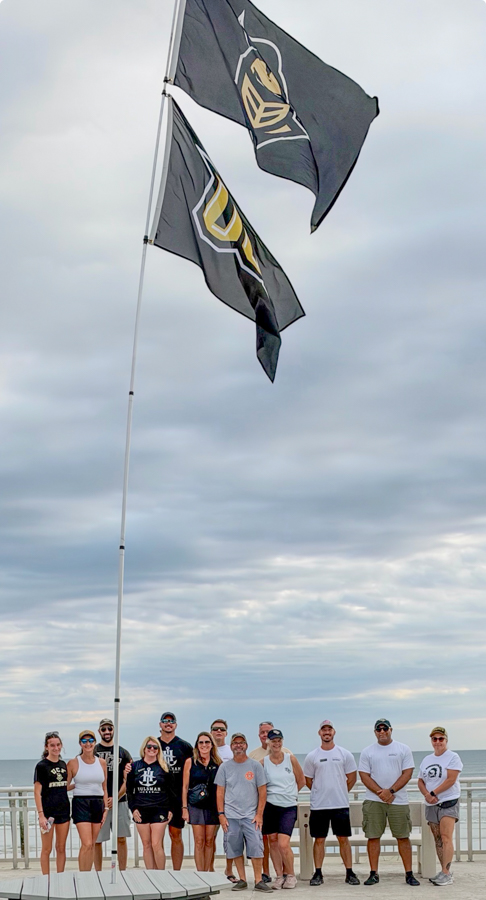 A group of people posing for a photo on a boardwalk by the ocean, with two tall flags waving above them.
