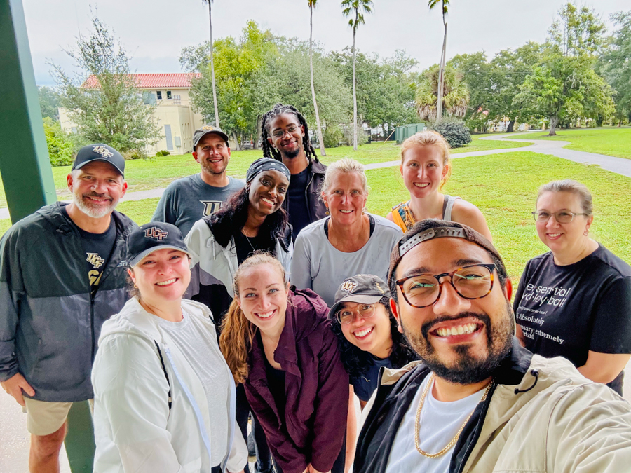 Group of adults posing for a selfie outdoors in a park, smiling and looking at the camera, with palm trees and green grass in the background.