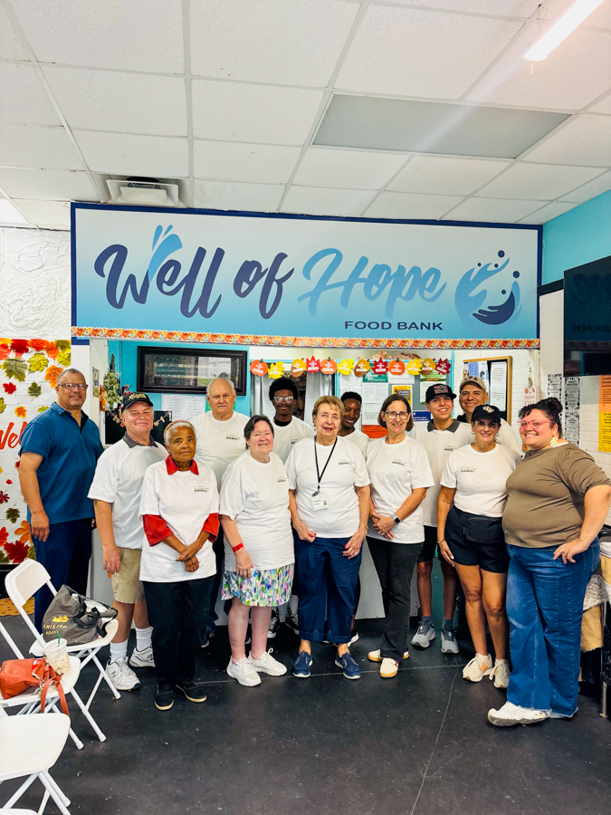Group of diverse volunteers standing together in front of a 'Well of Hope Food Bank' sign inside a community center.