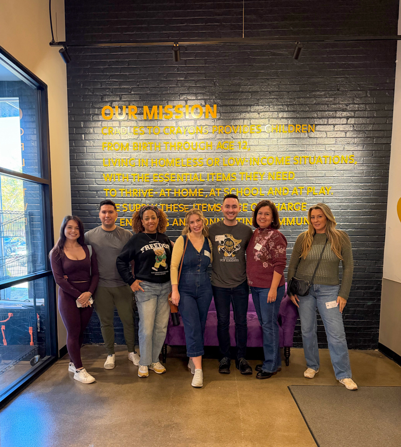 Eight people posing in a lobby in front of a black brick wall with a large yellow 'OUR MISSION' sign and text behind them.