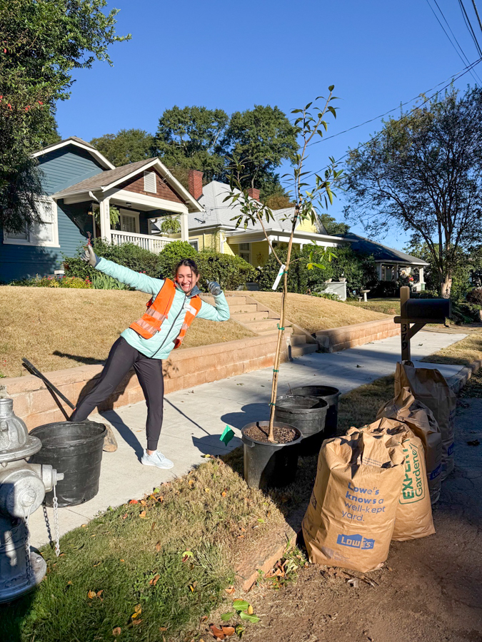 Woman in an orange safety vest posing with a newly planted sapling on a sunny suburban sidewalk, with planting bags and tools nearby.