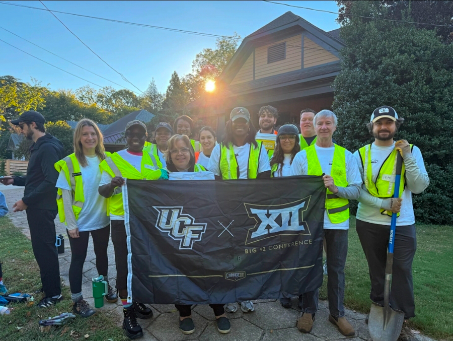 Group of volunteers in neon safety vests posing together while holding a banner that reads UCF × Big 12 Conference, outdoors in a suburban yard with a house in the background.