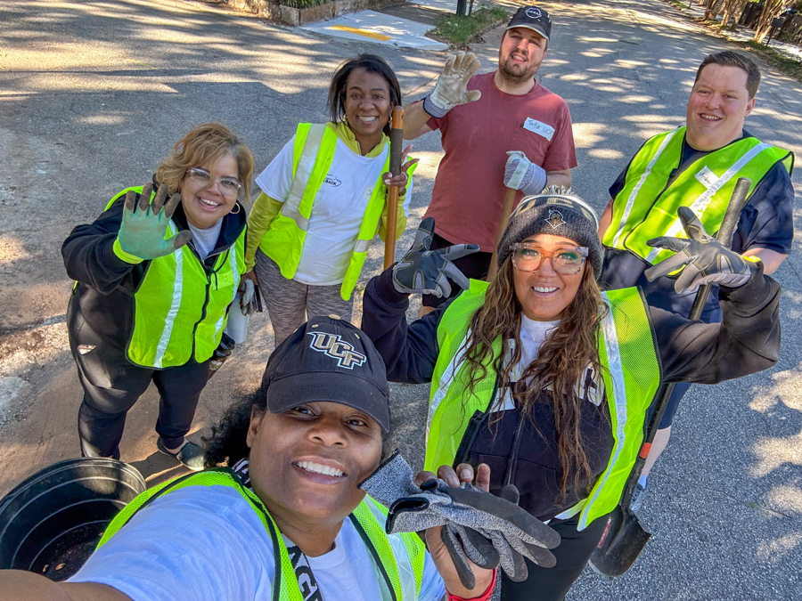Group of volunteers in safety vests waving at the camera during an outdoor cleanup.