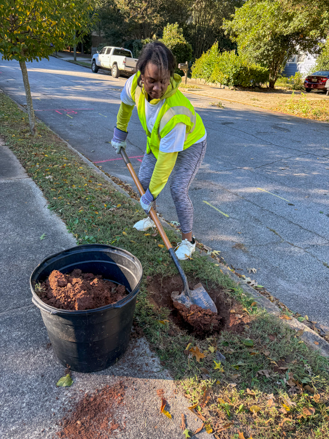 Person in a neon safety vest shoveling dirt from a bucket into a hole along the sidewalk during a community cleanup.