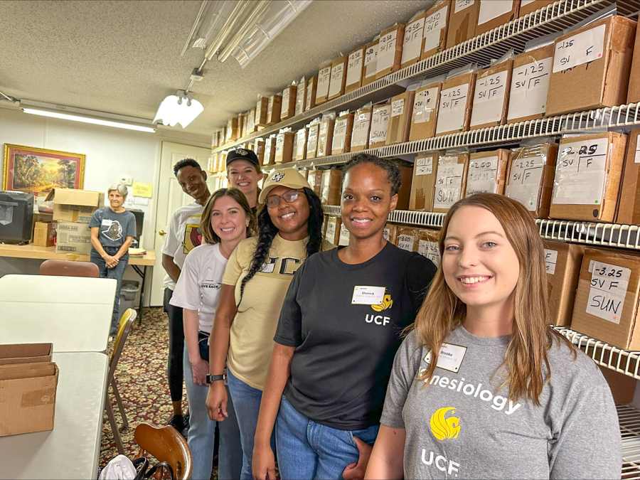 Seven volunteers pose in a storage room with labeled boxes on tall shelves, smiling at the camera.