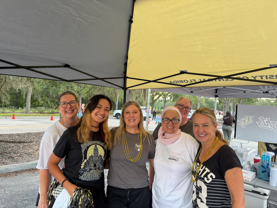 Six smiling women pose together under a yellow-and-gray canopy at an outdoor event, wearing casual clothes and bead necklaces for a group photo.